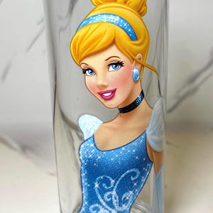 May include: A clear glass tumbler with a colourful illustration of Cinderella. The artwork depicts Cinderella's blonde updo, blue headband, and sparkling blue gown. The glass is cylindrical and is a standard drinking glass.
