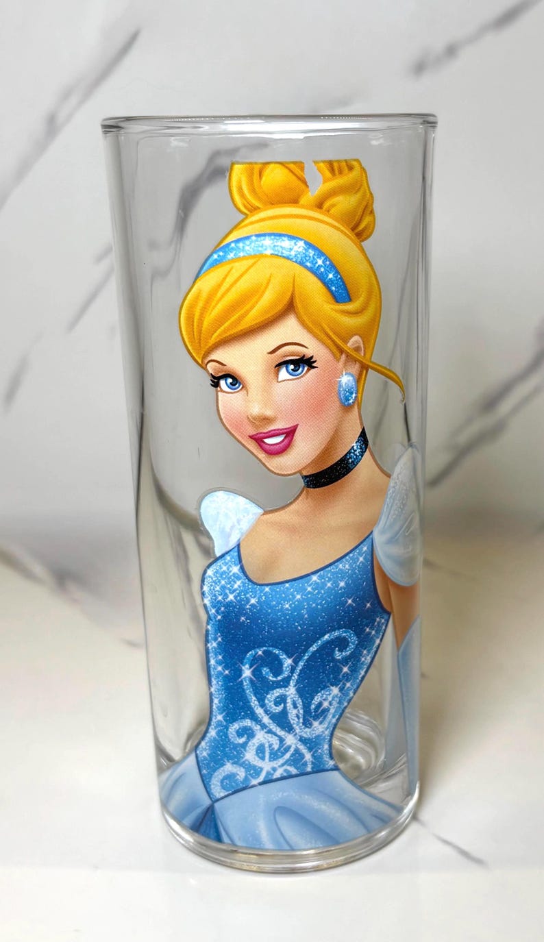 May include: A clear glass tumbler with a colourful illustration of Cinderella. The artwork depicts Cinderella's blonde updo, blue headband, and sparkling blue gown. The glass is cylindrical and is a standard drinking glass.