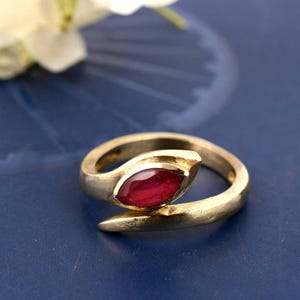 Peut inclure: Une bague dorée avec une pierre précieuse rouge foncé de taille marquise. La bague a un design ouvert unique avec une extrémité pointue. La bague est posée sur un fond bleu.