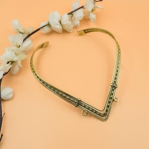 May include: A gold-colored collar necklace with a V-shaped design. The necklace has a series of small, decorative dots along the edges and a clasp closure. White flowers and branches are in the background.