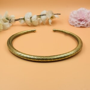 Puede incluir: Un collar dorado con un patrón floral detallado. El collar está abierto en la parte delantera y tiene extremos redondeados. El fondo es de color naranja claro con flores blancas y una flor rosa.