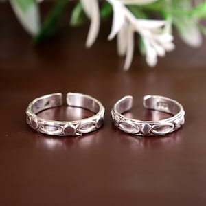 Puede incluir: Dos anillos de plata para el dedo gordo del pie con un diseño abierto. Cada anillo presenta una serie de elementos decorativos ovalados y redondos. Los anillos se muestran sobre una superficie marrón oscuro, con flores blancas en el fondo.