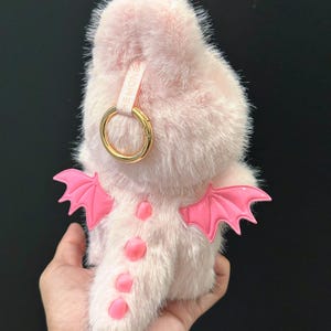 May include: A light pink, fluffy dragon-shaped accessory with bright pink wings and accents. It features a gold-colored ring and a small strap with text. The dragon has a soft, furry texture.