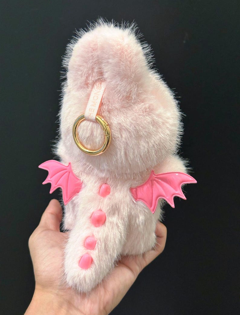 May include: A light pink, fluffy dragon-shaped accessory with bright pink wings and accents. It features a gold-colored ring and a small strap with text. The dragon has a soft, furry texture.