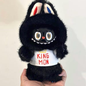 May include: A black plush toy figure with a bunny ear hood, wearing a white shirt with "KING MON" printed on it. The figure has large, round eyes and a small red nose. The bunny ears have red and blue accents.