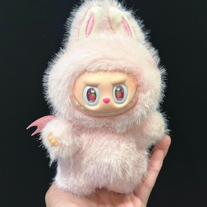 May include: A plush pink bunny-like figure with a smiling face and strawberry-like eyes. The figure has a furry exterior, small pink wings, and bunny ears. The figure is held in a hand against a dark background.