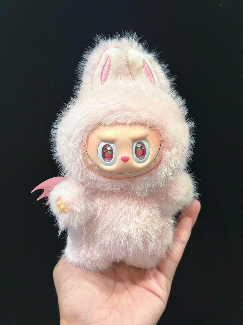 May include: A plush pink bunny-like figure with a smiling face and strawberry-like eyes. The figure has a furry exterior, small pink wings, and bunny ears. The figure is held in a hand against a dark background.