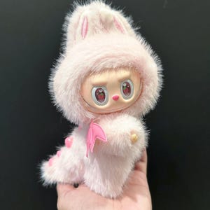 May include: A pink plush toy with a bunny-eared hood and a dragon-like tail. The toy has a doll-like face with strawberry-shaped eyes and a pink bat-wing accessory. The toy is held in a hand.