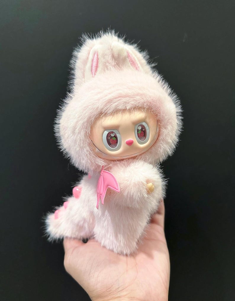May include: A pink plush toy with a bunny-eared hood and a dragon-like tail. The toy has a doll-like face with strawberry-shaped eyes and a pink bat-wing accessory. The toy is held in a hand.