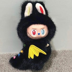 May include: A black plush toy in the shape of a character with a rabbit-like hood and a yellow wing. The character has a round face with mismatched red and blue eyes, and a small blue nose. The ears are white with yellow and red accents.