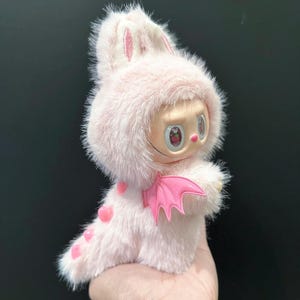 May include: A pink plush toy with a bunny-like hood and a small face with large eyes. The toy has pink heart-shaped spots and a pair of pink plastic wings. The toy is held in a hand.