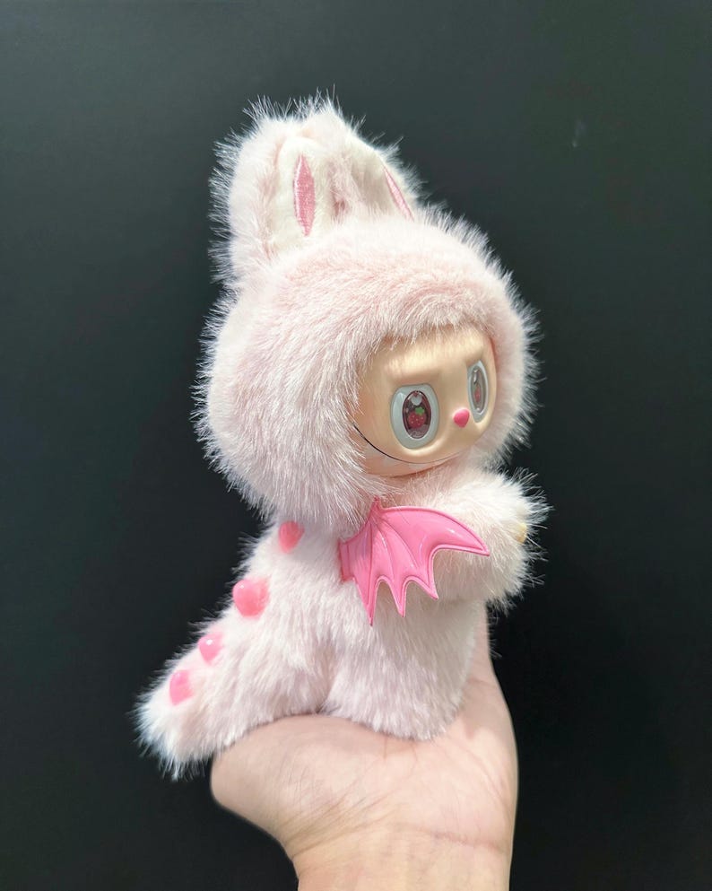 May include: A pink plush toy with a bunny-like hood and a small face with large eyes. The toy has pink heart-shaped spots and a pair of pink plastic wings. The toy is held in a hand.