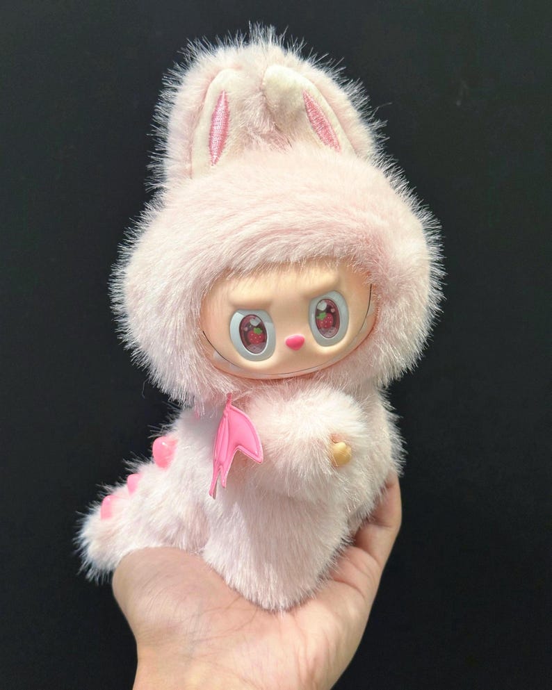 May include: A pink plush doll with a bunny hood and a small pink bow. The doll has a round face with large eyes and a small mouth. The doll's body is covered in soft, fluffy fur. The doll is being held in a hand.