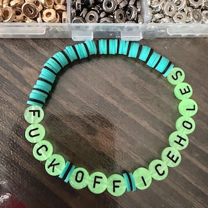 Peut inclure: Un bracelet fait à la main avec les mots "FUCK OFF ICE HOLES" épelés en lettres noires sur des perles vertes lumineuses. Le bracelet présente des sections alternées de perles en forme de disque turquoise et noires. L'arrière-plan comprend des conteneurs de petites perles métalliques.