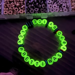 Peut inclure: Un bracelet de perles vert fluorescent avec les mots "ALEX PRETTI WAS ICY BY 87018" en lettres blanches. Le bracelet comporte des perles d'accentuation noires, bleues et rouges. L'arrière-plan montre des récipients de perles.