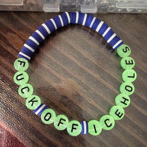 Peut inclure: Un bracelet de perles avec le texte "FUCK OFFICE HOLES" en lettres noires sur des perles vert clair. Le bracelet comporte une section de perles rayées bleues et blanches.