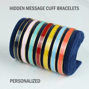 May include: A collection of eight metal cuff bracelets with colourful enamel inlay in black, pink, teal, red, yellow, light blue, and orange, displayed on a dark blue velvet holder. The text HIDDEN MESSAGE CUFF BRACELETS PERSONALIZED is visible.