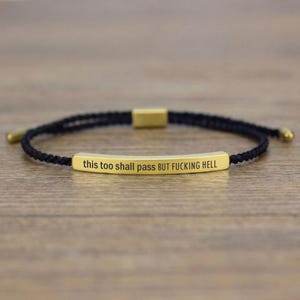May include: A black braided bracelet with a gold-colored rectangular bar in the center. The bar is engraved with the phrase "this too shall pass BUT FUCKING HELL". The bracelet has gold-colored end caps.
