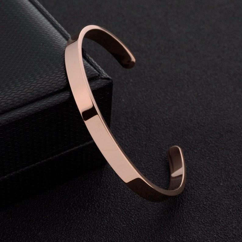 May include: A rose gold-toned metal cuff bracelet with a smooth, polished finish and a C-shaped open design. The bracelet features a flat, uniform band that curves into a circular shape, showcasing a reflective surface and clean, simple lines.
