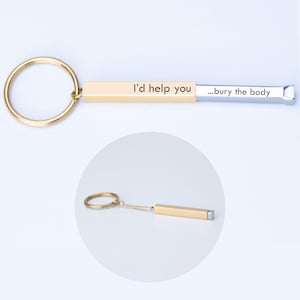 May include: A gold-colored keychain with a rectangular bar engraved with the text "I'd help you ...bury the body." The keychain has a gold ring and a small chain connecting the ring to the bar.