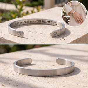 May include: A silver-toned metal cuff bracelet with a brushed finish. The interior is engraved with the text "this too shall pass BUT FUCKING HELL" and the ends are marked with small heart symbols. The bracelet is open-ended.