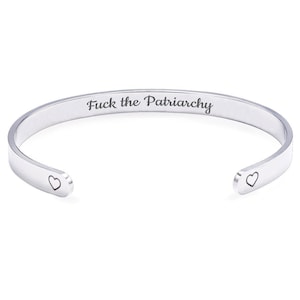 May include: A silver-colored cuff bracelet with the phrase "Fuck the Patriarchy" engraved on the outside. The bracelet has a heart-shaped design on each open end. The bracelet is made of a shiny metal.