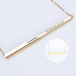 May include: A gold bar necklace featuring the phrases "remember" and "who the fuck you are". The pendant is rectangular and suspended from a fine chain. The design is minimalist and contemporary, suitable for daily wear.