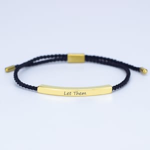 May include: A black cord bracelet featuring a gold-coloured rectangular bar engraved with the words "Let Them". The bracelet has gold-coloured end caps and an adjustable clasp. The cord is braided.