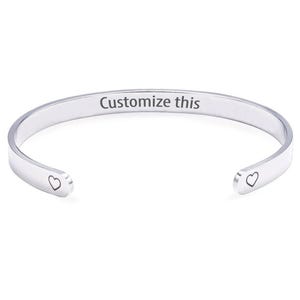 May include: A silver cuff bracelet with the text "Customize this" engraved inside. Each open end of the bracelet has a small heart design. The bracelet is designed for customisation. The metal is a bright, reflective silver.