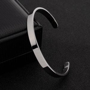 May include: A polished silver-tone metal cuff bracelet with a smooth, flat surface and a C-shaped open design. The bracelet features a reflective finish and rounded edges, showcasing a clean, simple aesthetic without any additional ornamentation.