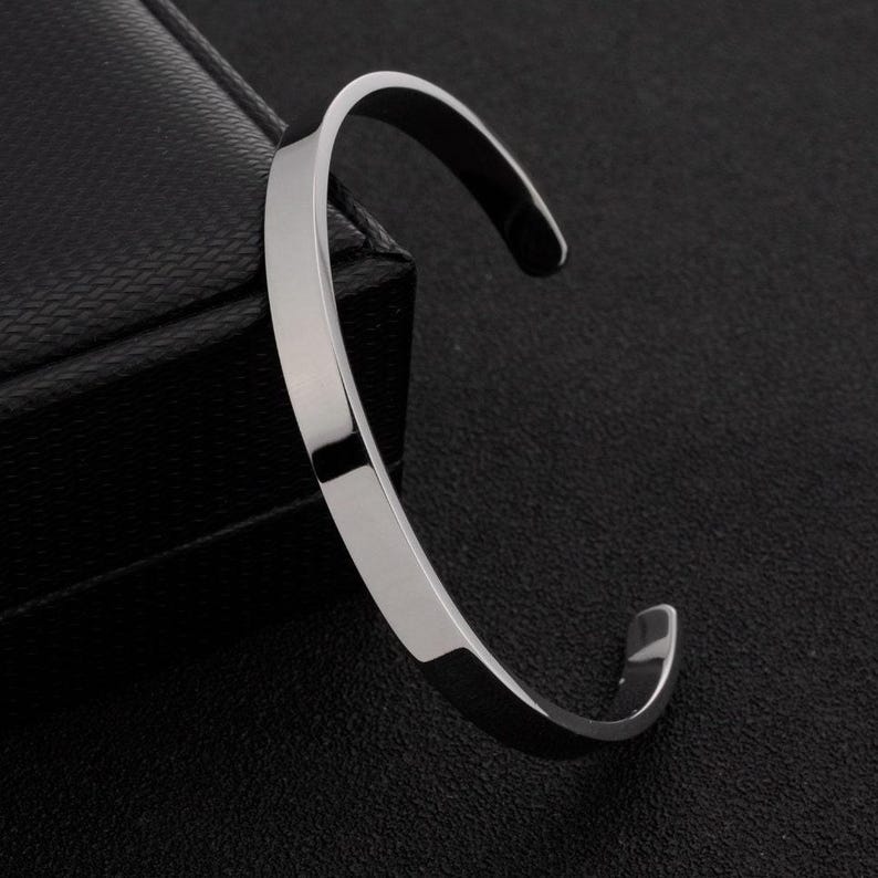 May include: A polished silver-tone metal cuff bracelet with a smooth, flat surface and a C-shaped open design. The bracelet features a reflective finish and rounded edges, showcasing a clean, simple aesthetic without any additional ornamentation.