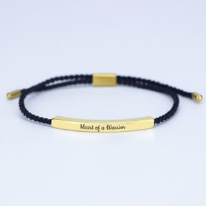 May include: A black braided cord bracelet with gold-colored metal accents. The bracelet features a rectangular gold bar engraved with the words "Heart of a Warrior". The bracelet is adjustable.