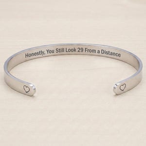 May include: A silver-toned metal cuff bracelet with an open design. The outer surface is engraved with the text "Honestly, You Still Look 29 From a Distance" and features a small heart symbol at each of the two rounded ends of the bracelet.