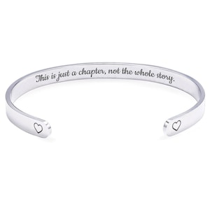 May include: A silver cuff bracelet with the engraved text "This is just a chapter, not the whole story." The bracelet features a heart-shaped design on each open end.