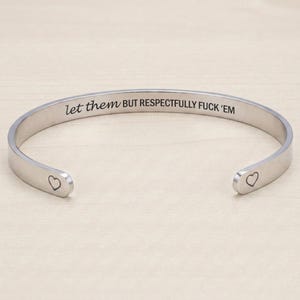 May include: A silver-toned metal cuff bracelet featuring the engraved text 'let them BUT RESPECTFULLY FUCK 'EM' along the inner band, with a small heart symbol engraved at each of the two open ends of the bracelet.