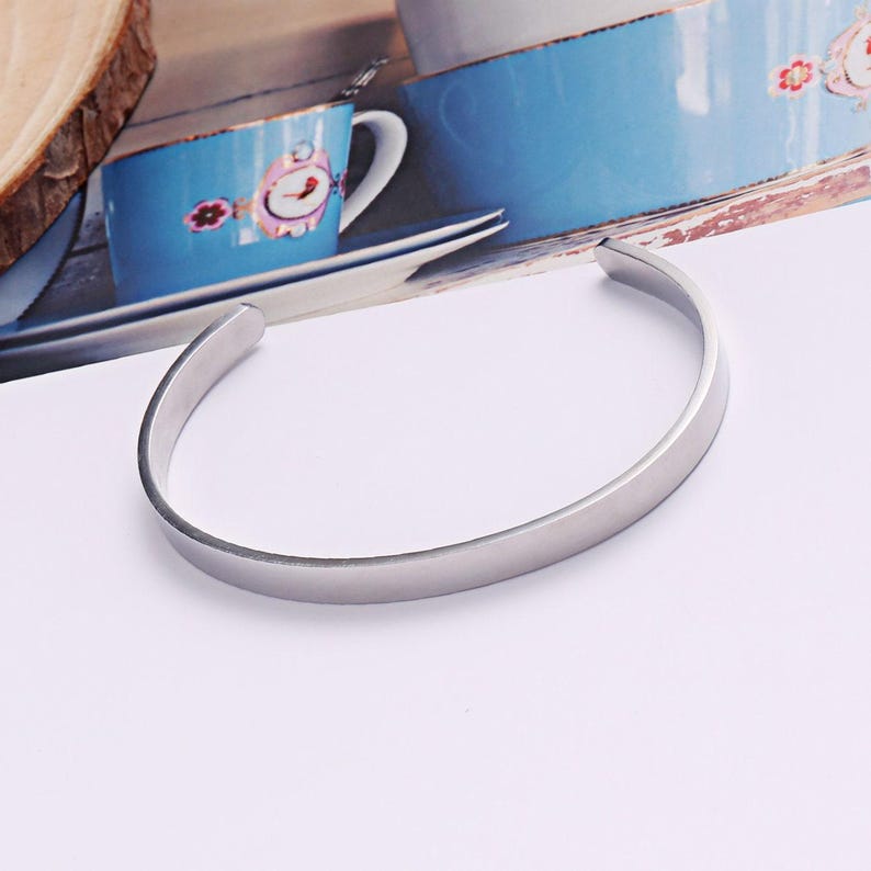 May include: A silver-toned metal cuff bracelet with a smooth, polished finish and a simple open-ended design. The bracelet features a rounded, minimalist band that curves into a circular shape, showcasing a clean and reflective metallic surface.
