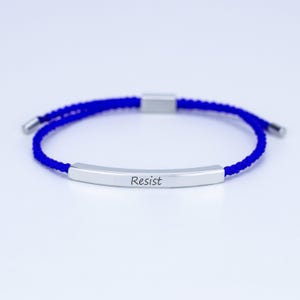 May include: A blue adjustable bracelet with a silver bar engraved with the word "Resist". The bracelet features a woven cord and silver end caps. A simple, yet powerful statement piece.