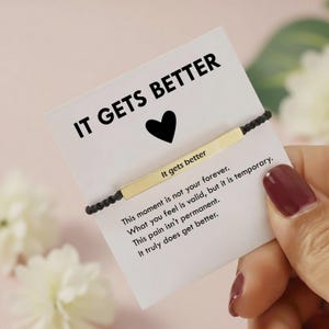 May include: A gold bar bracelet with the words "It gets better" on a white card with the text "IT GETS BETTER" and a black heart. The card also includes the text "This moment is not your forever. What you feel is valid, but it is temporary."