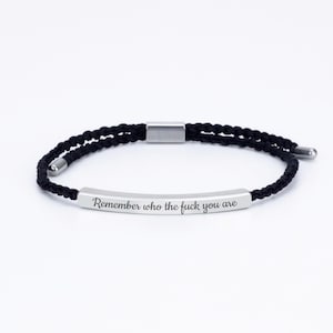 Peut inclure: Bracelet tressé noir avec une barre argentée gravée de la phrase « Remember who the fuck you are ». Le bracelet a un design minimaliste et une fermeture à fermoir. Le bracelet est en cordon noir.