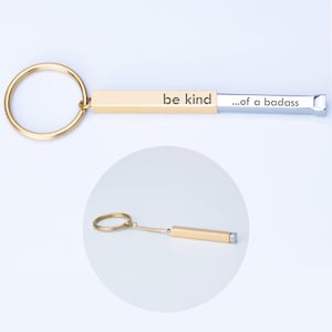 May include: A gold-coloured keychain with the words "be kind" and "...of a badass" printed on it. The keychain has a gold ring and a rectangular bar. The background is white.