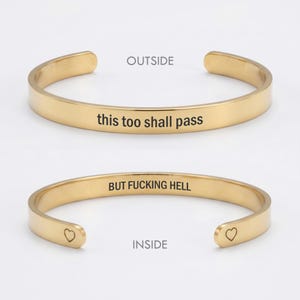 May include: A gold-toned metal cuff bracelet shown from two angles. The outside is engraved with the text 'this too shall pass'. The inside is engraved with the text 'BUT FUCKING HELL' and features a small heart symbol at each open end.