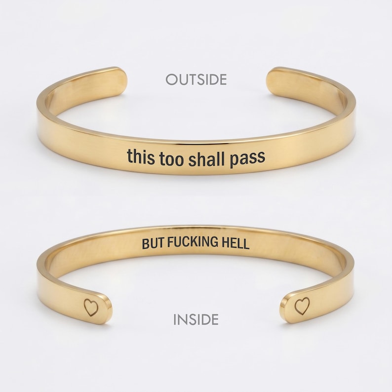 May include: A gold-toned metal cuff bracelet shown from two angles. The outside is engraved with the text 'this too shall pass'. The inside is engraved with the text 'BUT FUCKING HELL' and features a small heart symbol at each open end.