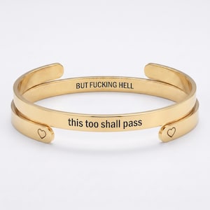 May include: A gold-toned metal cuff bracelet featuring an open design. The outer surface is engraved with the text "this too shall pass" and small heart symbols at the ends. The inner surface is engraved with the text "BUT FUCKING HELL".