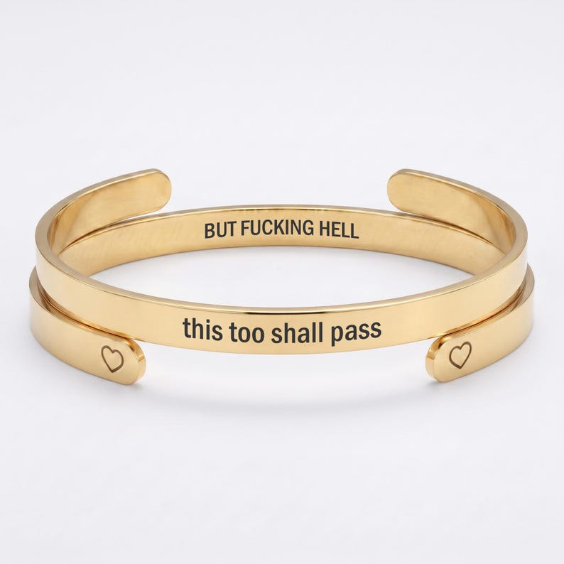 May include: A gold-toned metal cuff bracelet featuring an open design. The outer surface is engraved with the text "this too shall pass" and small heart symbols at the ends. The inner surface is engraved with the text "BUT FUCKING HELL".
