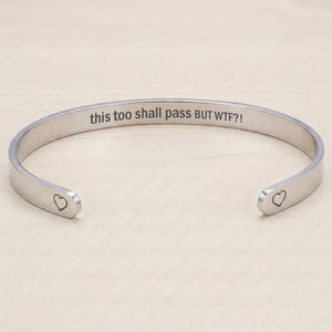 Peut inclure: Un bracelet jonc en métal argenté avec une finition polie. L'intérieur est gravé avec le texte "this too shall pass BUT WTF?!" et chaque extrémité arrondie du bracelet présente un petit contour simple en forme de cœur.