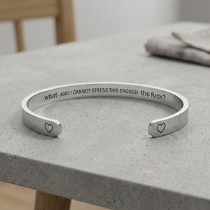 May include: A silver cuff bracelet with the engraved text "what -AND I CANNOT STRESS THIS ENOUGH- the fuck?" and a heart symbol on each end. The bracelet is displayed on a grey surface.