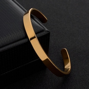 May include: A gold-toned metal cuff bracelet with a smooth, polished finish and a simple open-ended design. The bracelet features a flat, uniform band that curves into a circular shape, showcasing a reflective surface that catches the light.