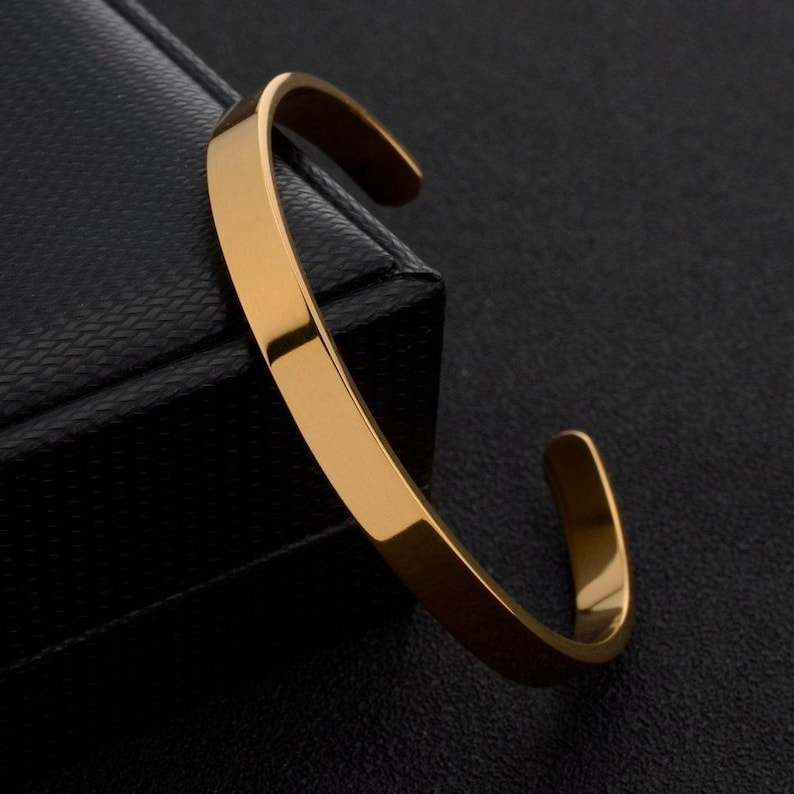 May include: A gold-toned metal cuff bracelet with a smooth, polished finish and a simple open-ended design. The bracelet features a flat, uniform band that curves into a circular shape, showcasing a reflective surface that catches the light.