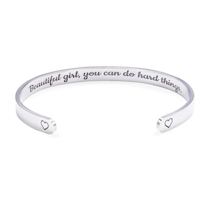 May include: A silver-colored cuff bracelet with the engraved text "Beautiful girl, you can do hard things." Small heart-shaped designs are at each open end. The bracelet is a simple, elegant design.