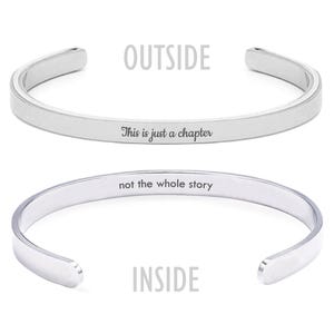 May include: A silver cuff bracelet with the text "This is just a chapter" engraved on the outside and "not the whole story" on the inside. The bracelet is open-ended and has a smooth, polished finish.